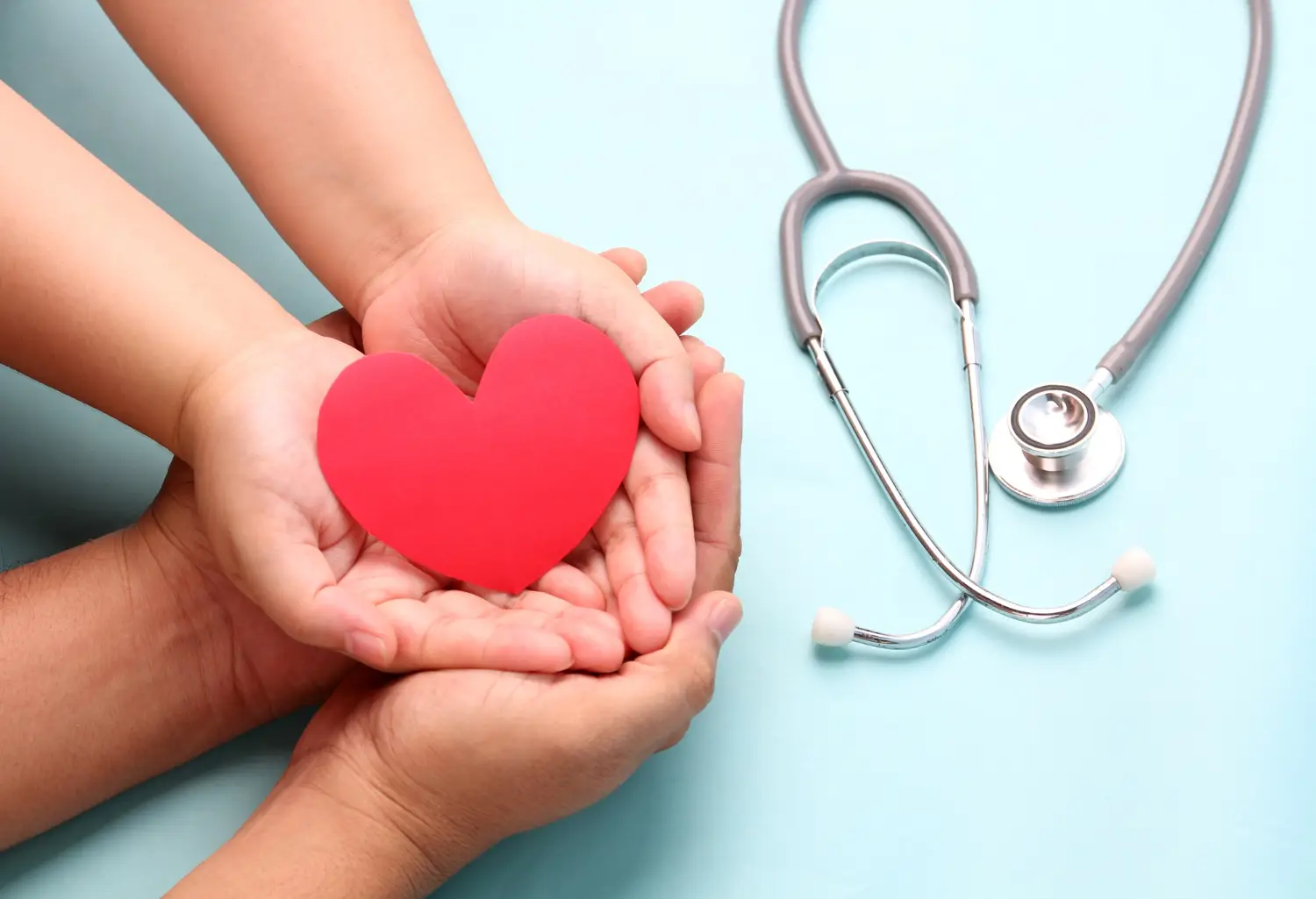 Hands holding paper red heart with stethoscope on blue.